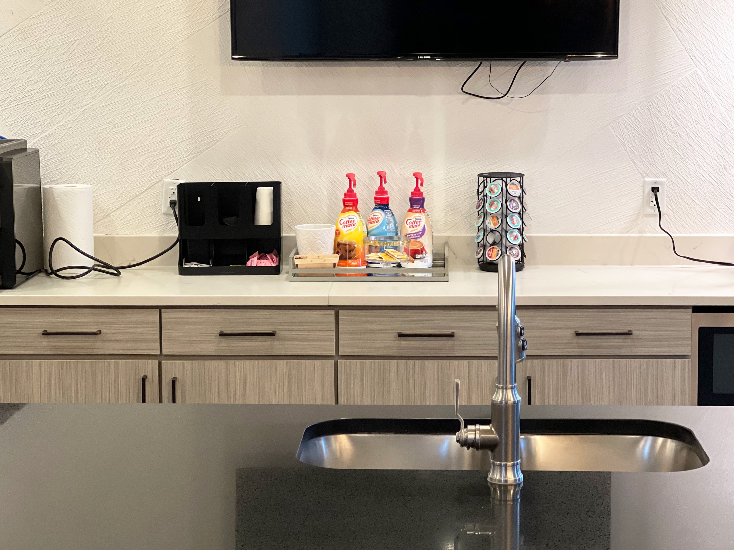 Coffee station with three pump bottles of coffee mate flavored creamers, a cup, a coffee stirer, and a selection of coffee capsules on a spice rack.