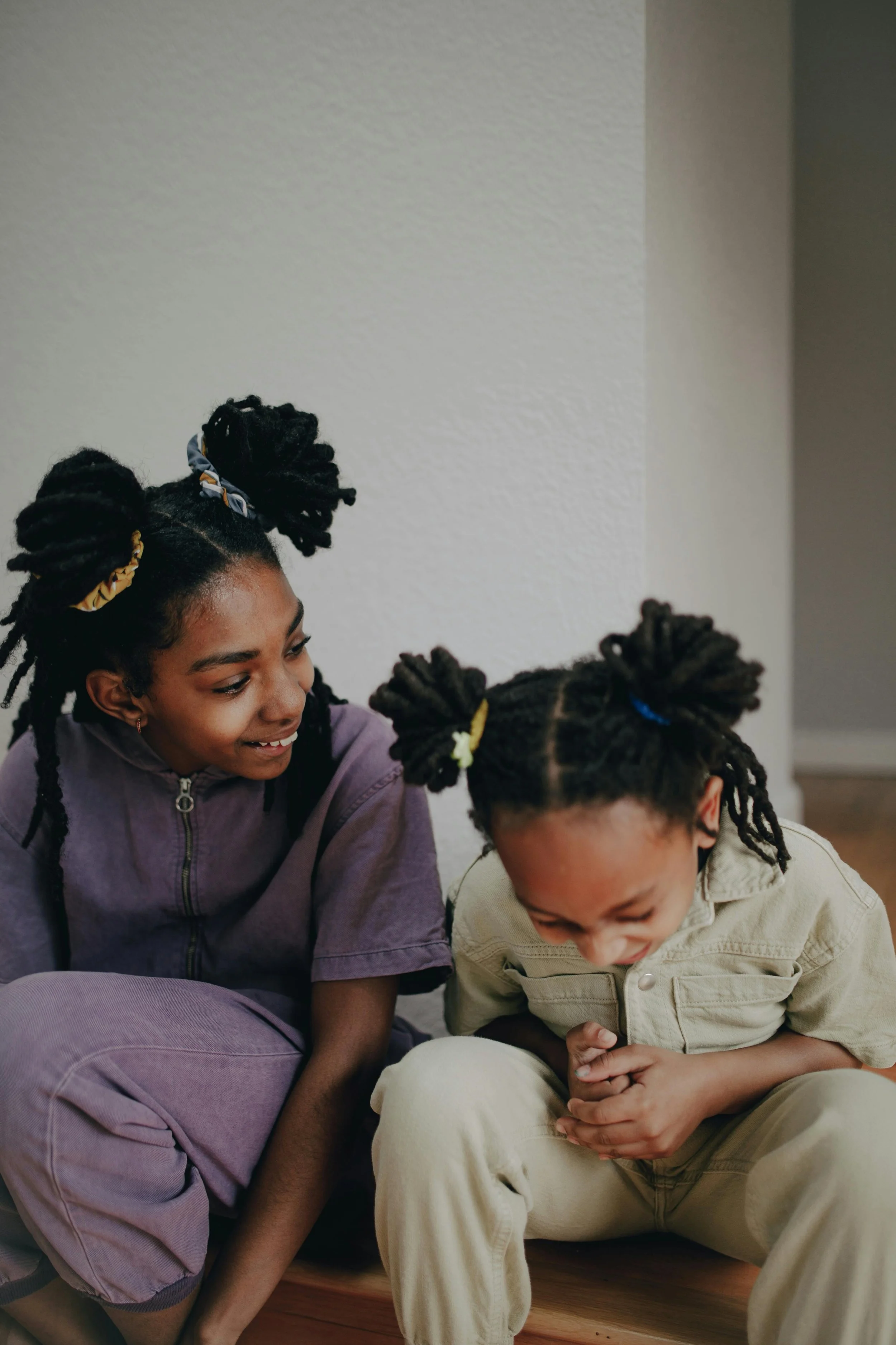 Two young girls with twisted hair in bun hairstyles, smiling and laughing while sitting on a wooden floor in a room with white walls.
