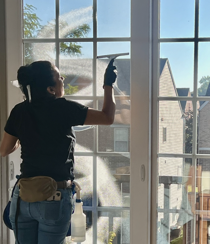 A woman in black shirt and gloves cleaning glass door with a spray bottle and cloth, sunlight streaming in.