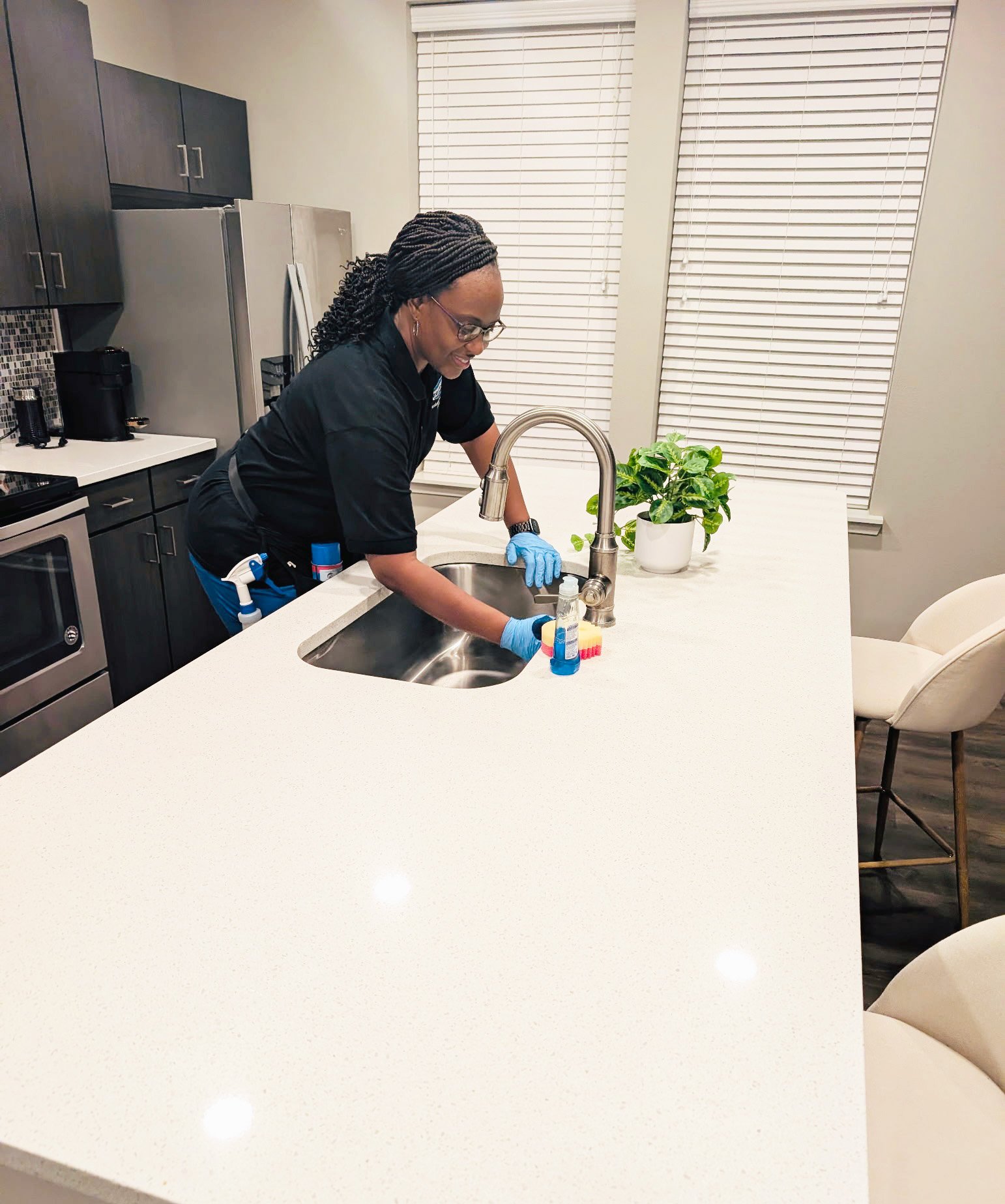 Woman cleaning a kitchen sink wearing gloves, with cleaning supplies nearby, in a modern kitchen with white countertops and dark cabinets.