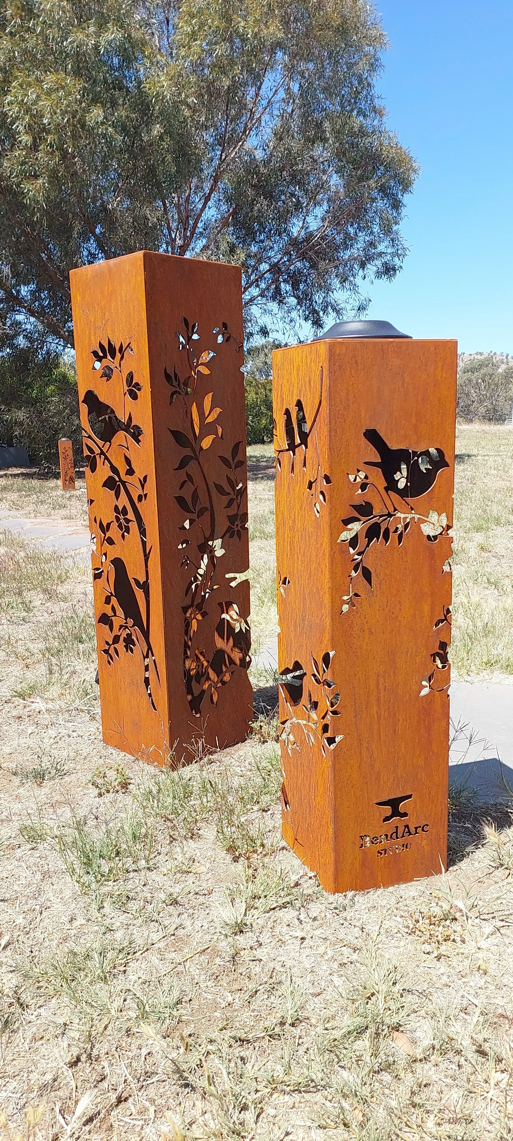 Two outdoor rust-colored metal sculptures with cutout designs of birds and leaves, set in a grassy area with trees in the background.