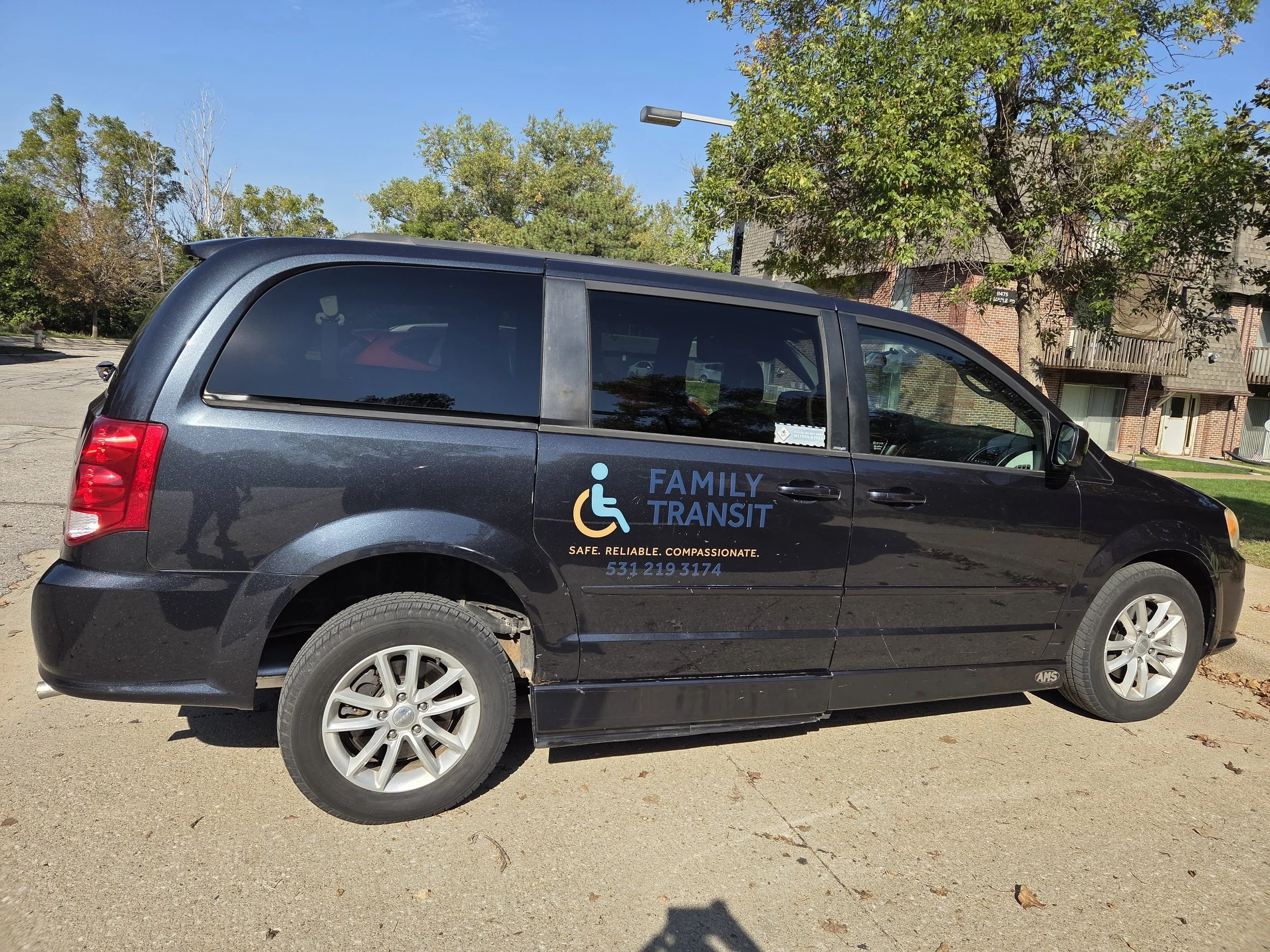 Black family transit minivan with accessible logo and contact information parked on a street with trees and residential buildings in the background.