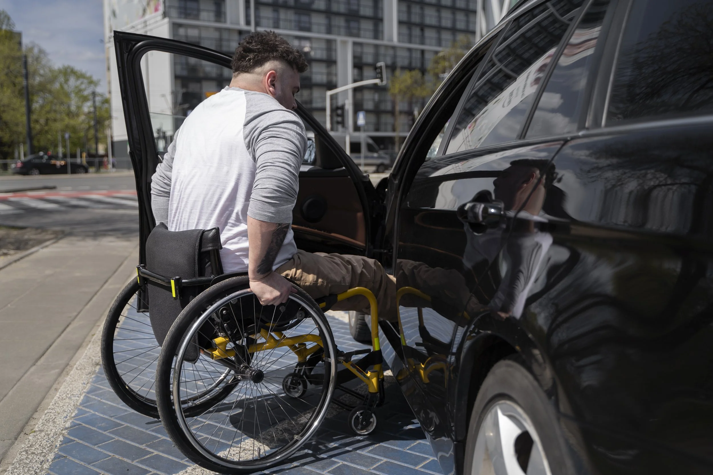A man in a wheelchair entering a black vehicle with the help of a person inside the car, on a city street during daytime.
