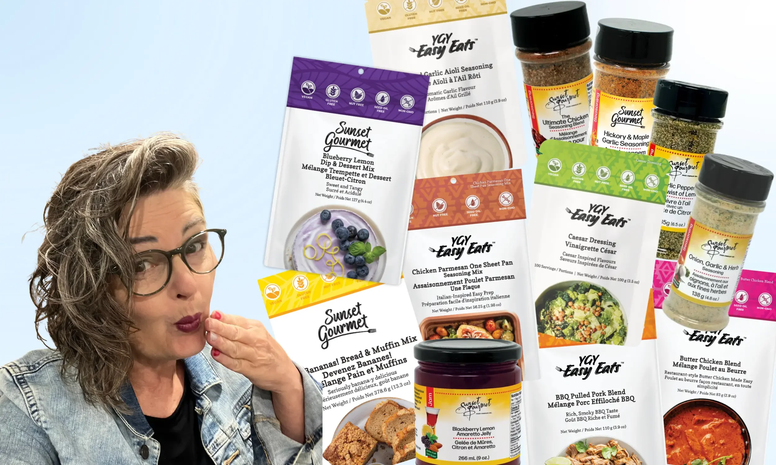A woman with brown curly hair, glasses, and a denim jacket is holding a small red object near her mouth as if about to eat or taste it. She is surrounded by various packets of seasoning and seasoning blends from the brand Sunset Gourmet, with flavors like blueberry lemon dip, garlic aioli, chicken parmesan, and BBQ pulled pork. Some bowls of food are also visible beneath the seasoning packets.