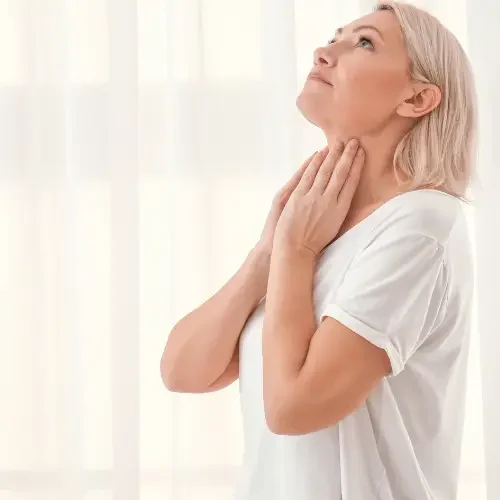 An elderly woman with gray hair touching her neck and looking upward in a well-lit room curious about her Thyroid.