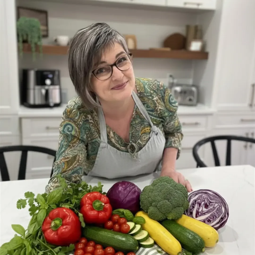 A woman wearing glasses and a patterned blouse with a gray apron, smiling and leaning over a white kitchen counter with a variety of fresh vegetables including red bell peppers, tomatoes, cucumbers, zucchini, broccoli, red cabbage, and herbs.