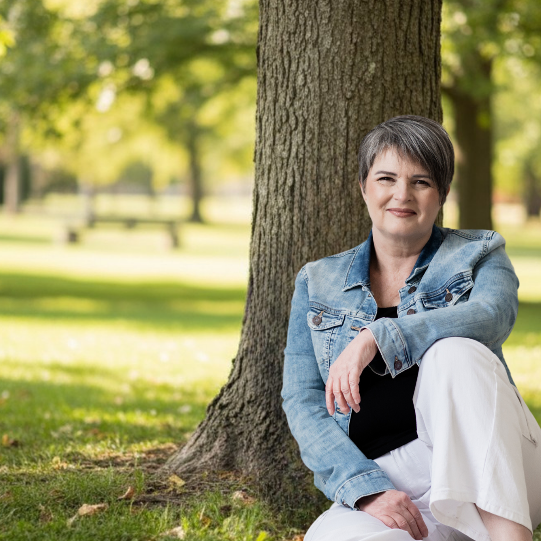 A woman with short gray hair, wearing a denim jacket and white pants, sitting on the grass near a tree in a park, smiling at the camera.