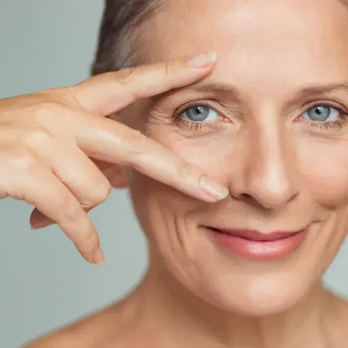 A woman with light skin and blue eyes holds her right hand near her eye, making a peace sign with her fingers.