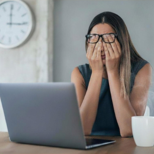 Woman with glasses covering face with hands sitting at a desk with laptop and coffee cup, clock on wall in background.