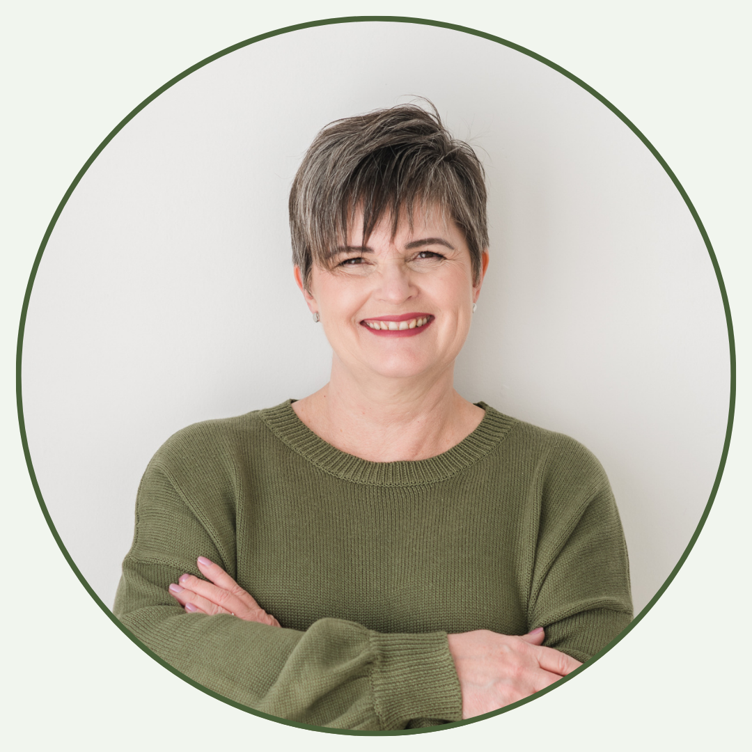 A smiling woman with short brown hair, wearing a green sweater, standing with arms crossed against a plain white background.