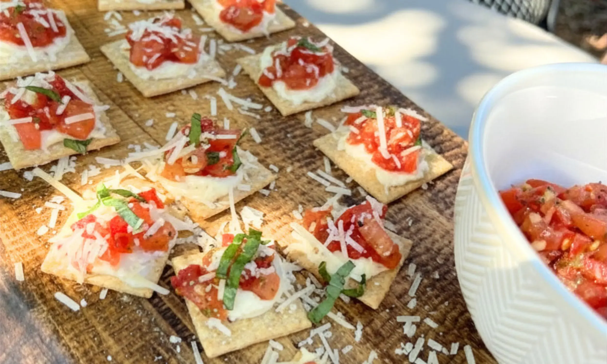 Miniature appetizers topped with tomato, basil, cheese, served on small crackers, with shredded cheese on a wooden surface and a bowl of tomato topping on the side.