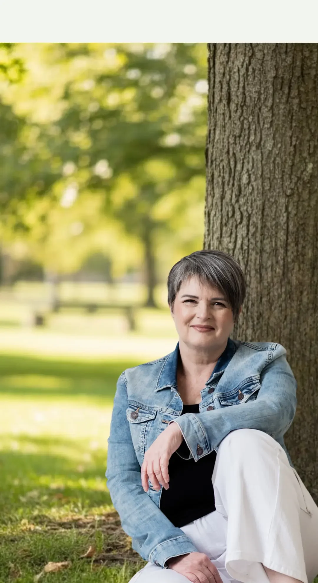 A woman with short gray hair, wearing a denim jacket and white pants, sitting outdoors near a tree in a park with green grass and blurred trees in the background.