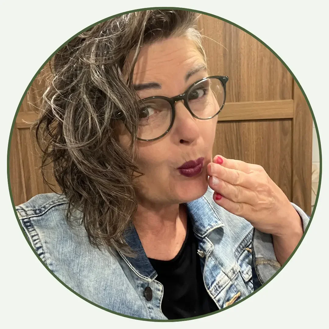 A woman with short, curly hair and glasses, wearing a denim jacket, touching her lips with her hand, standing against a wooden background.