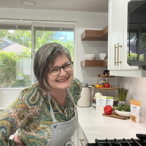 Smiling woman with gray hair and glasses wearing a patterned shirt and white apron in a modern kitchen with white cabinets, open shelves, and fresh vegetables on the counter.
