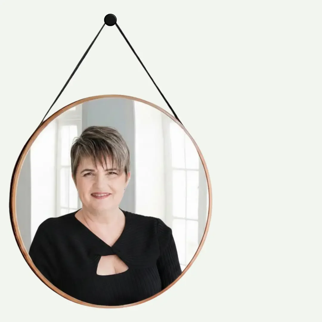 A mirror with a wooden frame hanging on a wall, reflecting a smiling woman with short brown hair wearing a black top, standing in front of windows with white curtains.