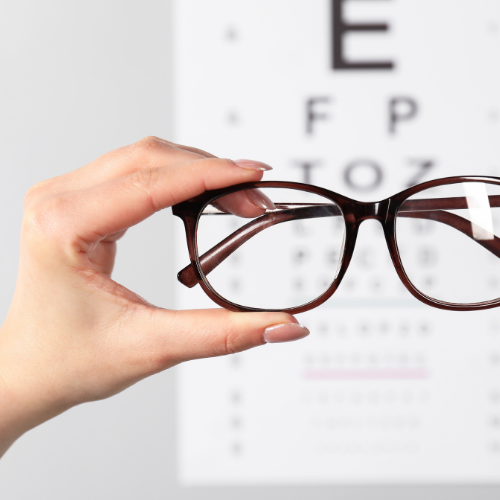 A person's hand holding a pair of eyeglasses in front of an eye chart.