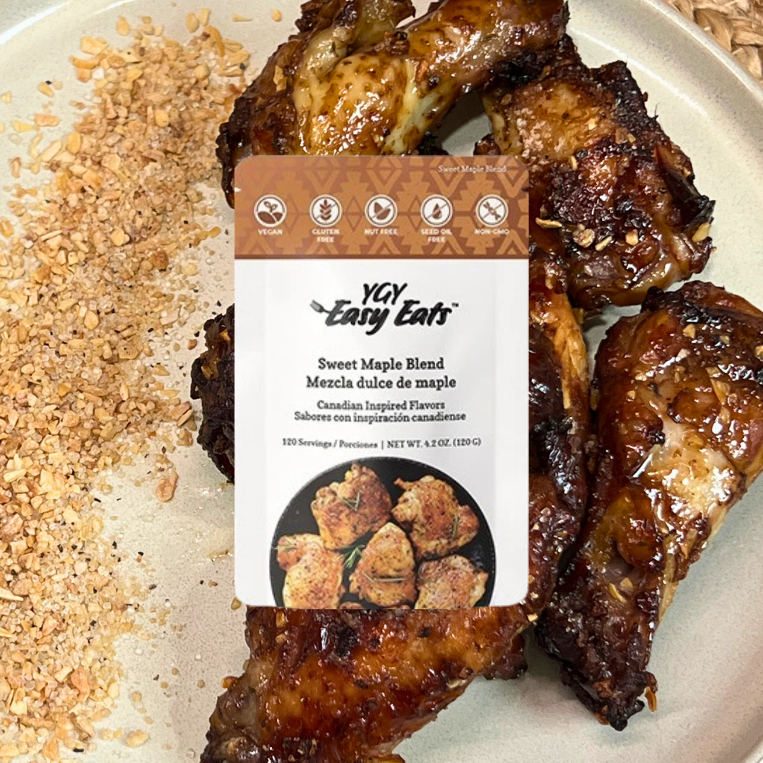Five glazed chicken drumsticks with a dark caramelized glaze on a plate, with chopped nuts or breadcrumbs sprinkled beside them, and a package of Sweet Maple Blend seasoned coating from Yay Easy Eats resting on the drumsticks.