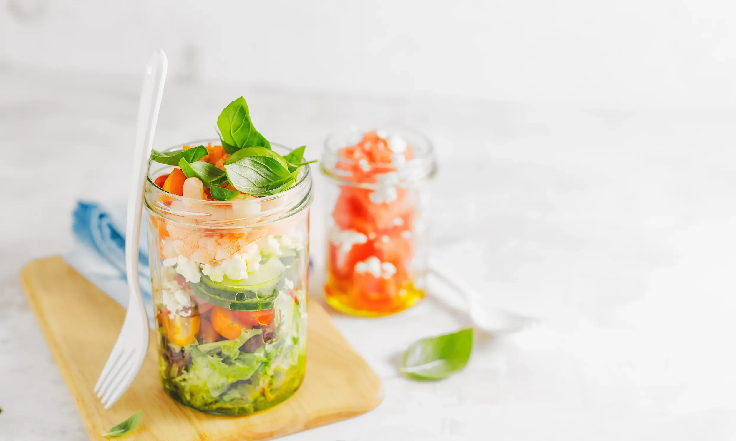 A tall glass jar filled with layered fresh vegetables and herbs, topped with basil leaves, with a fork leaning against it, on a wooden cutting board with another jar of chopped vegetables in the background on a white surface.