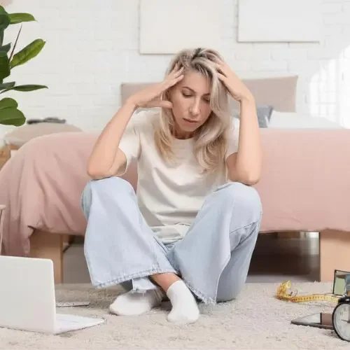 A woman sitting on the floor with a stressed expression, her hands on her head, surrounded by a laptop, measuring tape, and a clock in a bedroom.