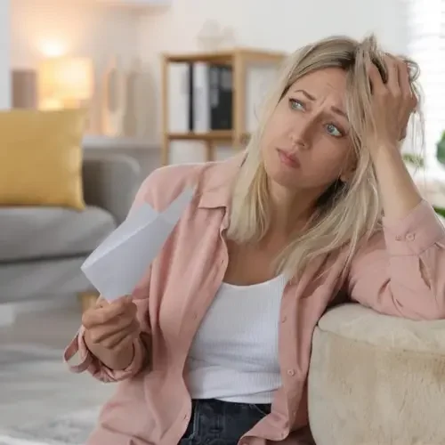A woman seated on a sofa, holding a letter and looking puzzled or confused while scratching her head.