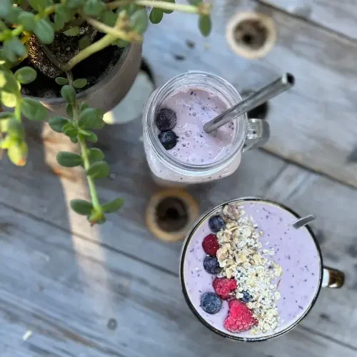Two smoothies for health protein morning starts  topped with berries and oats on a wooden table next to a potted plant.