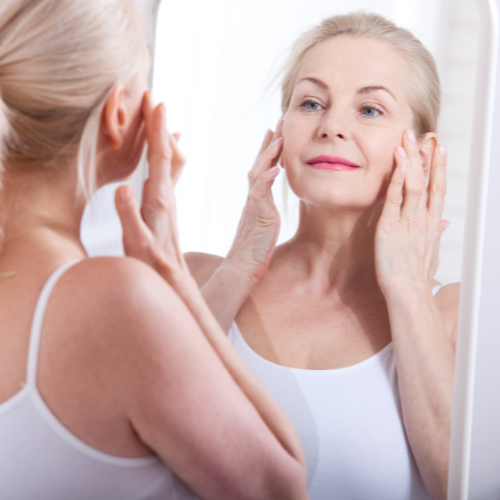 An older woman looking at herself in a mirror, touching her face with her hands, with natural light coming in
