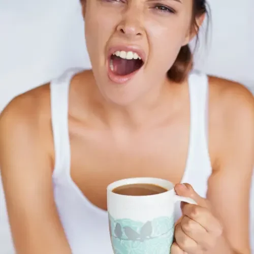 A woman in a white tank top is holding a mug of coffee or tea and appears to be yawning or in discomfort.