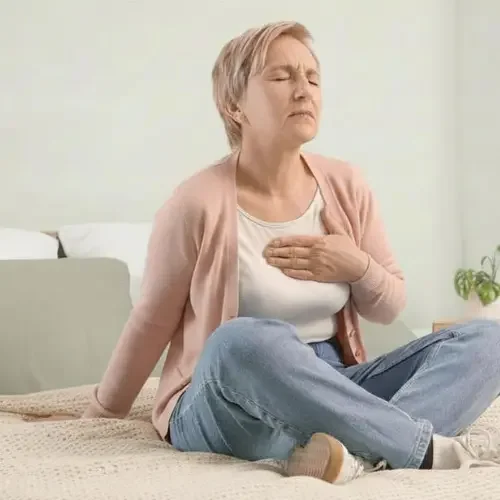 An older woman sitting on a bed, clutching her chest with a pained expression, indicating she may be experiencing chest pain or discomfort.
