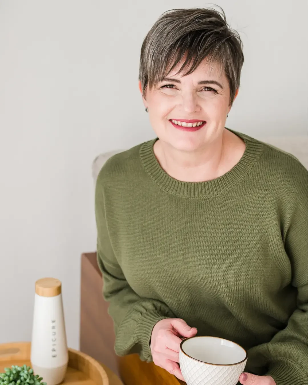A woman with short brown hair, smiling, wearing an olive green sweater, holding a white mug with a gold rim, sitting at a table with a small plant and a paper towel roll in a room with a neutral background.