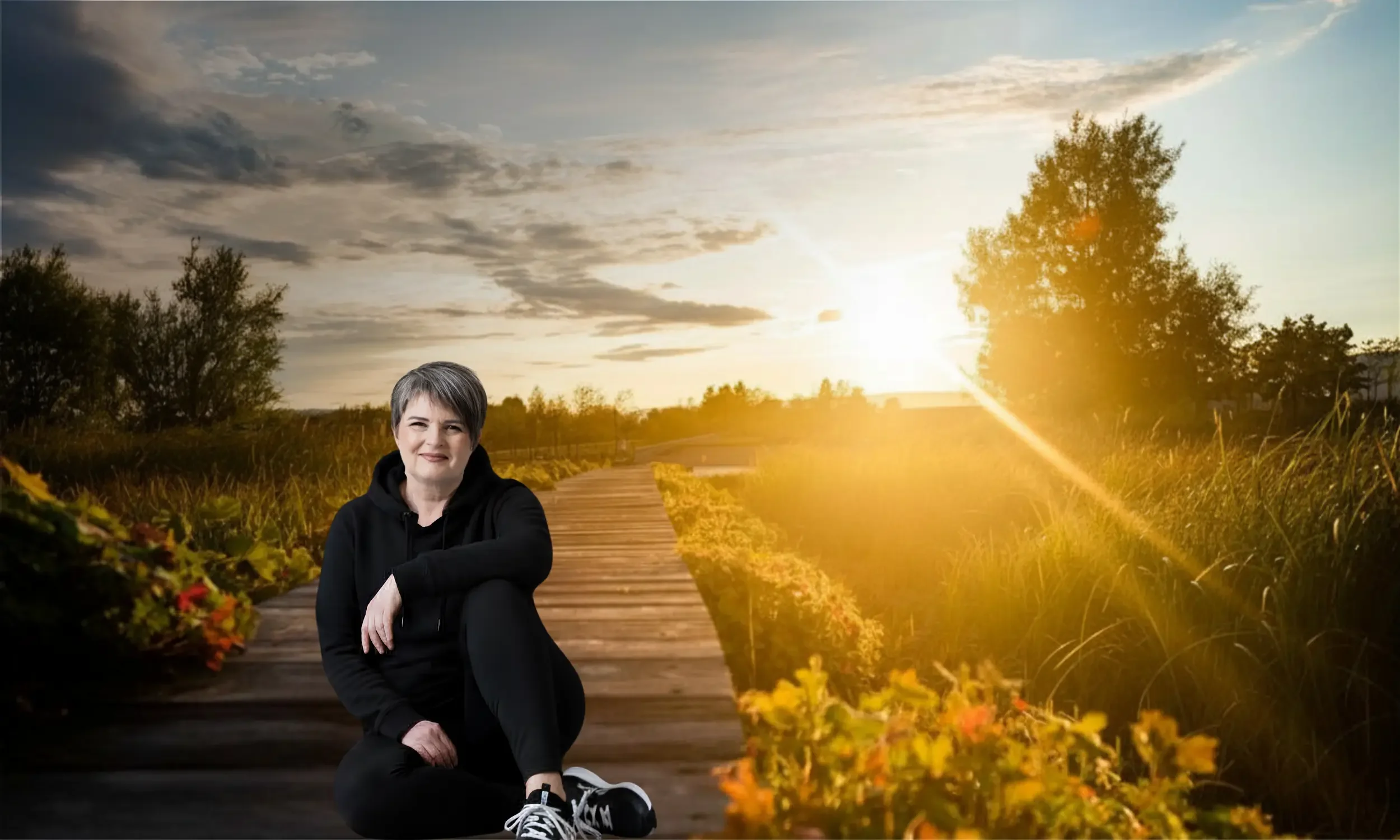 A digital composite image featuring a woman with short gray hair dressed in a black hoodie and black pants, sitting on a wooden pathway surrounded by yellow and orange flowers. The background displays a scenic landscape with a sunset, trees, and a partly cloudy sky.