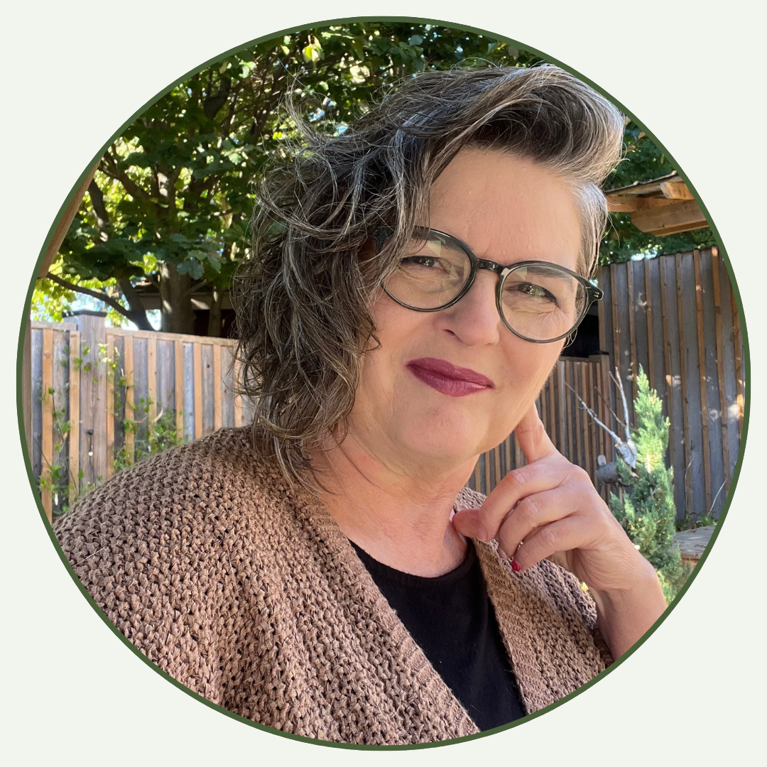 A woman with short, curly hair and glasses, wearing a brown sweater, posing outdoors near a wooden fence and green trees.