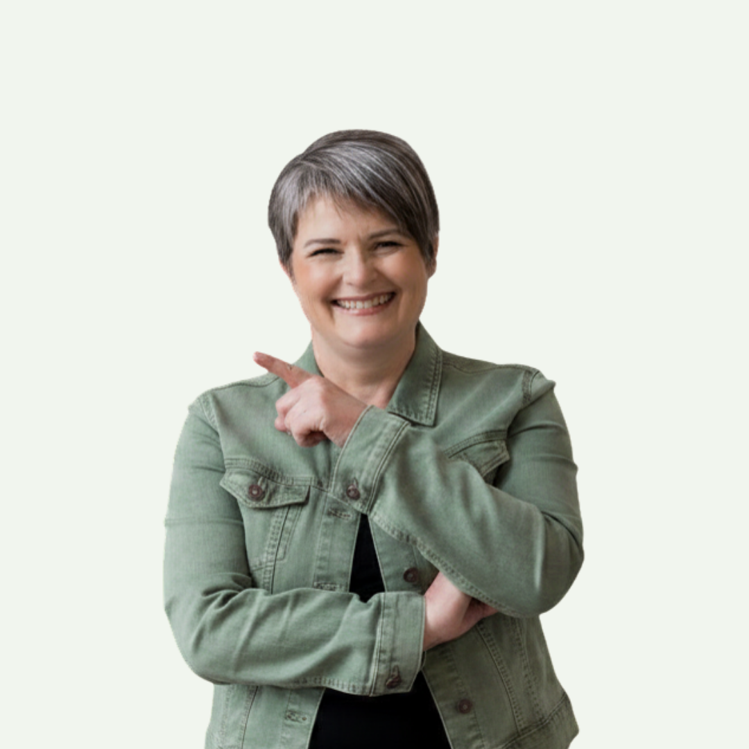 A smiling woman with short gray hair wearing a green denim jacket pointing her right index finger to the side against a plain light background.