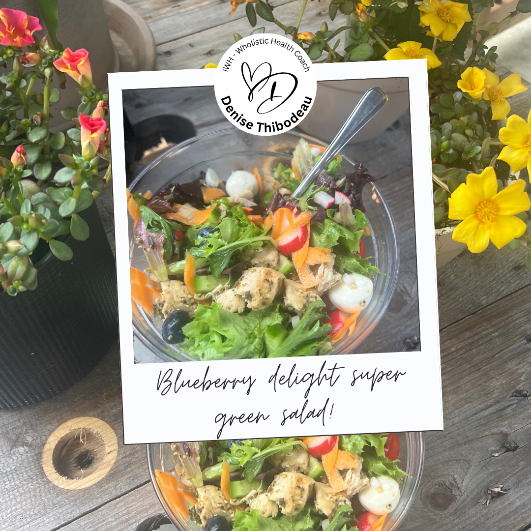 A bowl of Blueberry Delight Super Green Salad with various greens, blueberries, chopped vegetables, and chicken, placed on a wooden table decorated with potted plants and flowers.