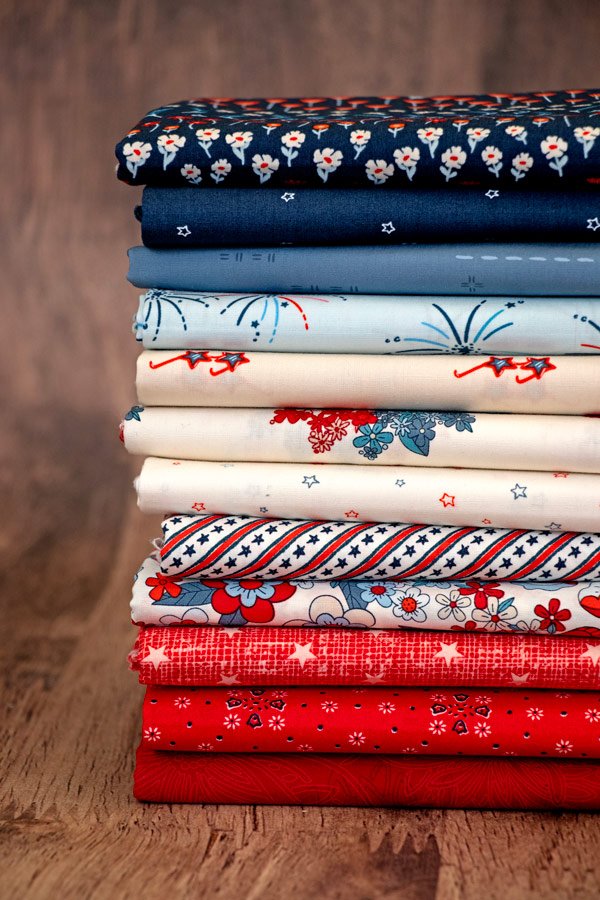 Stacked fabric with blue, white, and red floral and patriotic patterns on a wooden surface.