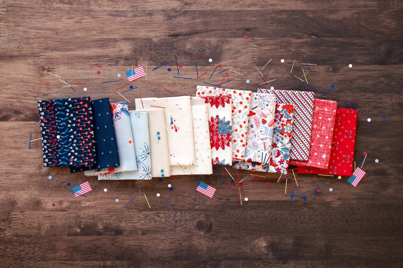 A collection of folded patriotic-themed fabric pieces in red, white, and blue, arranged on a wooden surface with scattered confetti, toothpicks, and small American flags.
