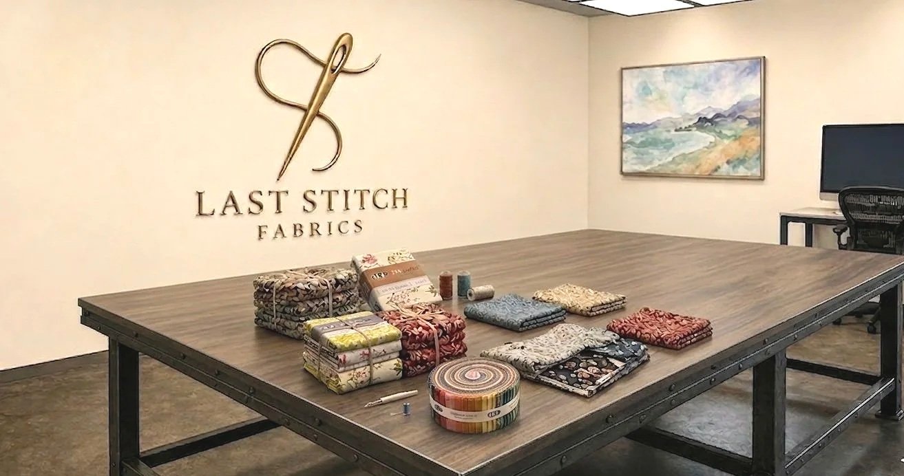 Fabric samples and bolts arranged on a large table with a sign for Last Stitch Fabrics on the wall.