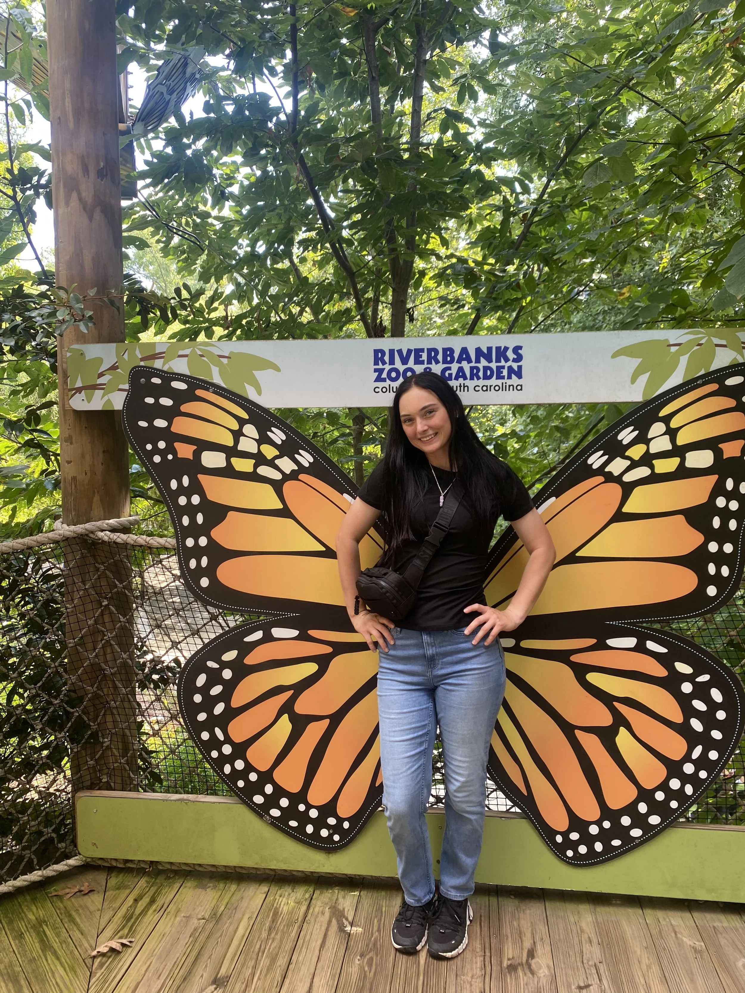 A young woman standing in front of a large butterfly wing display at Riverbanks Zoo Garden in South Carolina. She is smiling, wearing a black t-shirt and jeans, with a crossbody bag. Surrounding greenery and trees are visible in the background.