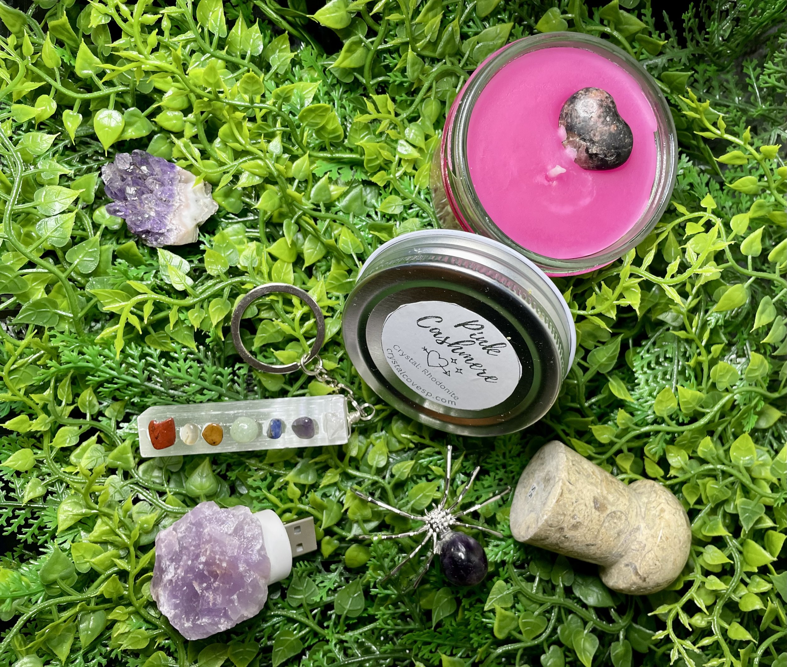 A collection of crystals, stones, candles, and jewelry arranged on green artificial plants. Items include a pink candle with a stone on top, a purple geode USB flash drive, a keychain with five small colorful stones, a labeled brown crystal, and a large polished stone.