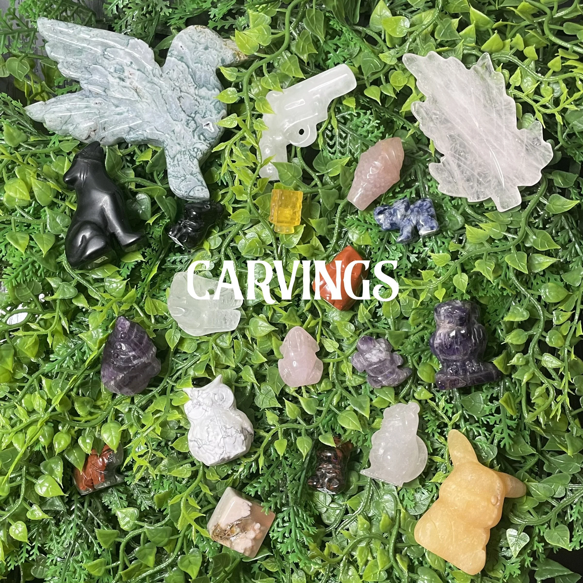 A collection of carved gemstone animals and objects placed on green artificial plants, including a bird, bear, cat, rabbit, owl, and various small carved stones, with the word 'CARVINGS' overlaid in white text.