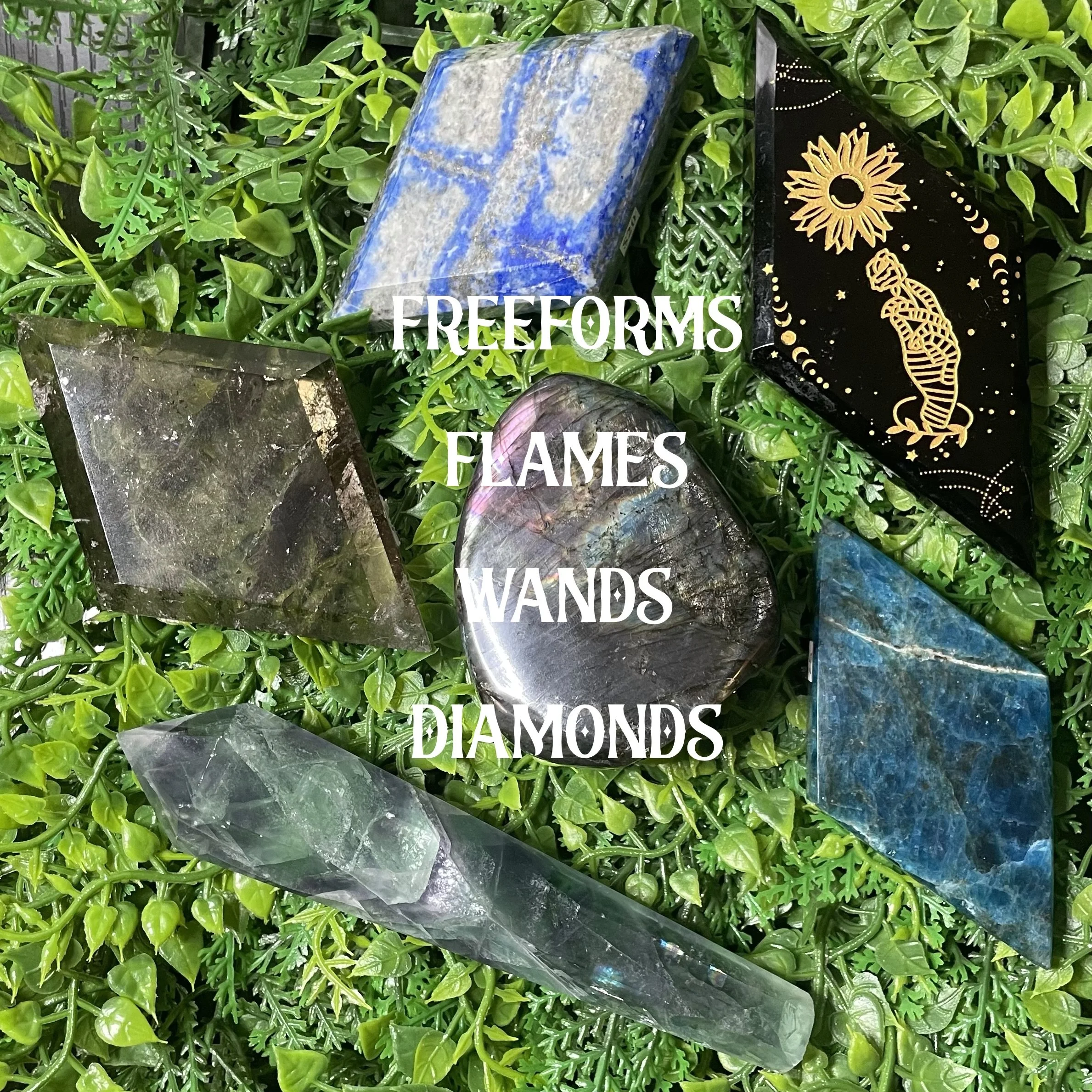 Several crystals and stones, including a heart-shaped labradorite, a black onyx with gold mystical designs, a tall clear quartz point, and a blue lapis lazuli, arranged on green foliage. Text overlay reads 'Freeforms, Flames, Wands, Diamonds.'