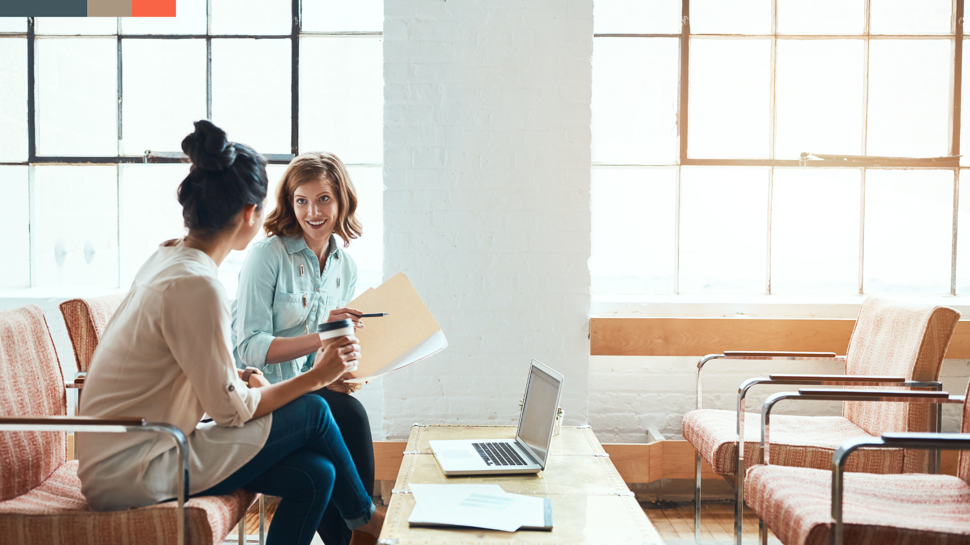 Two women having a conversation in a bright, modern room with large industrial-style windows. One woman is holding a coffee cup and some papers, while the other is smiling and talking. A laptop is open on a table between them.