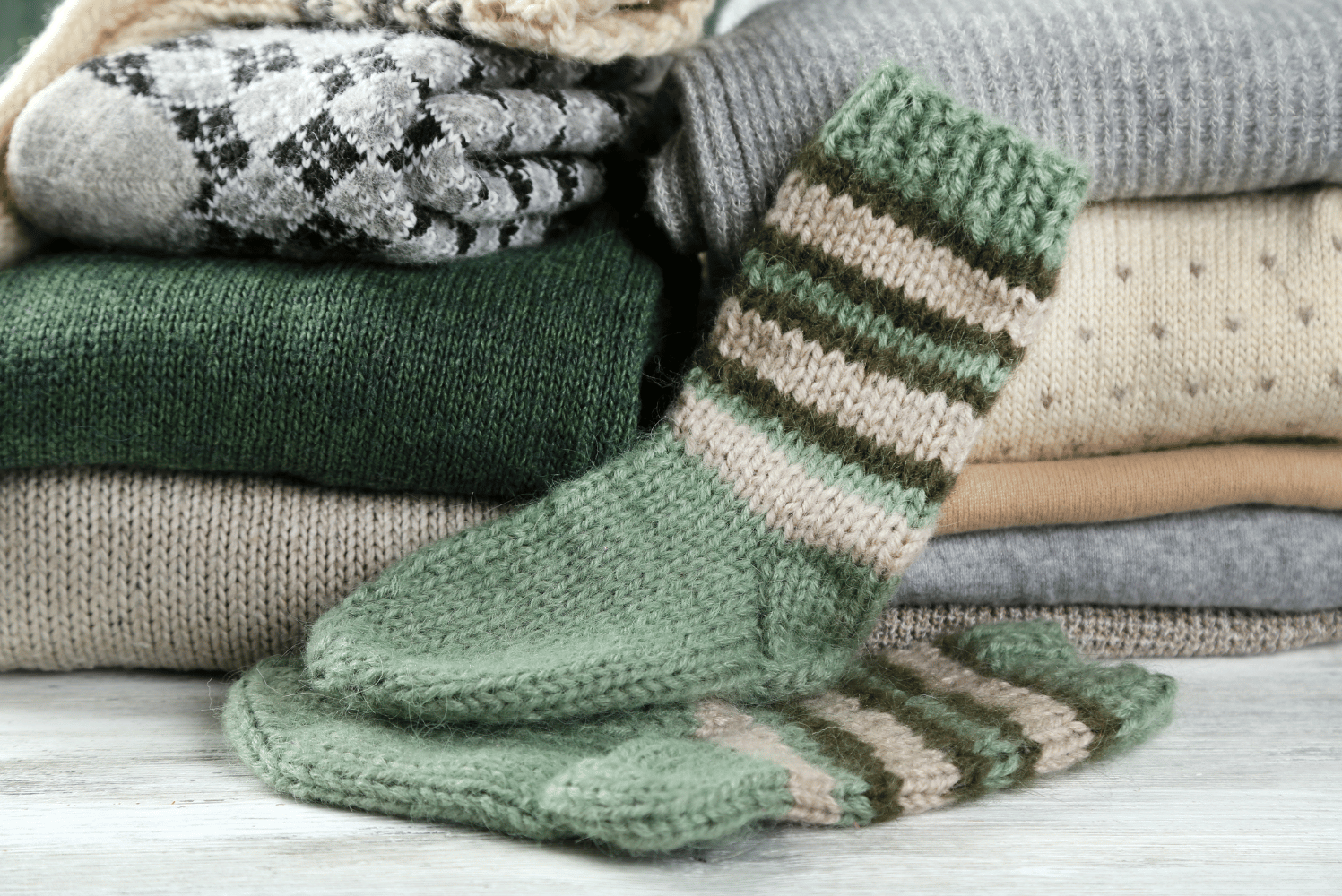 Stacked folded sweaters in neutral and earth tones with a green and beige striped knit sock draped over the side.