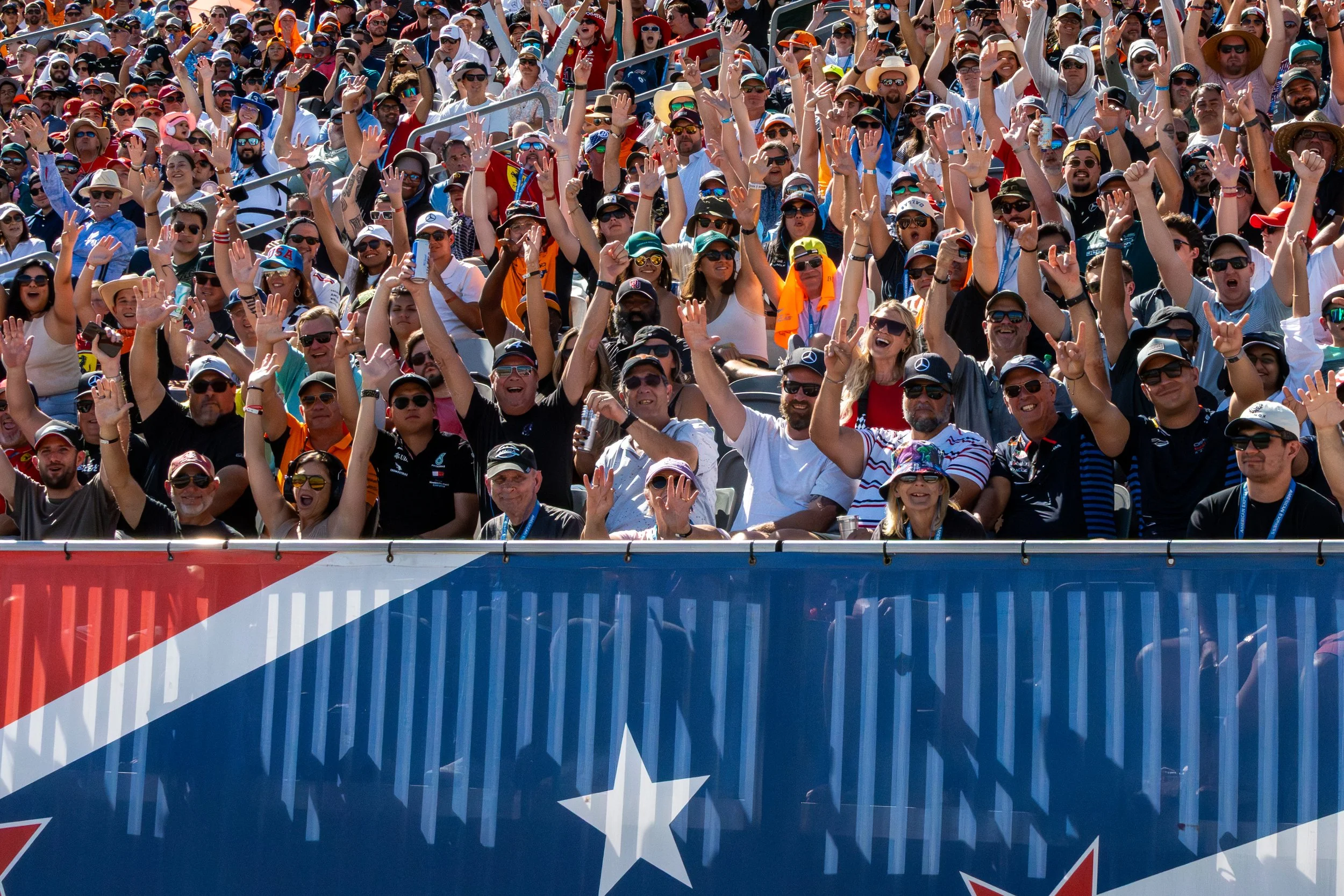 Crowd of people at a race event cheering, waving, and wearing sunglasses and hats, with a barrier or fence in the foreground and orange tents and a sunny sky in the background.