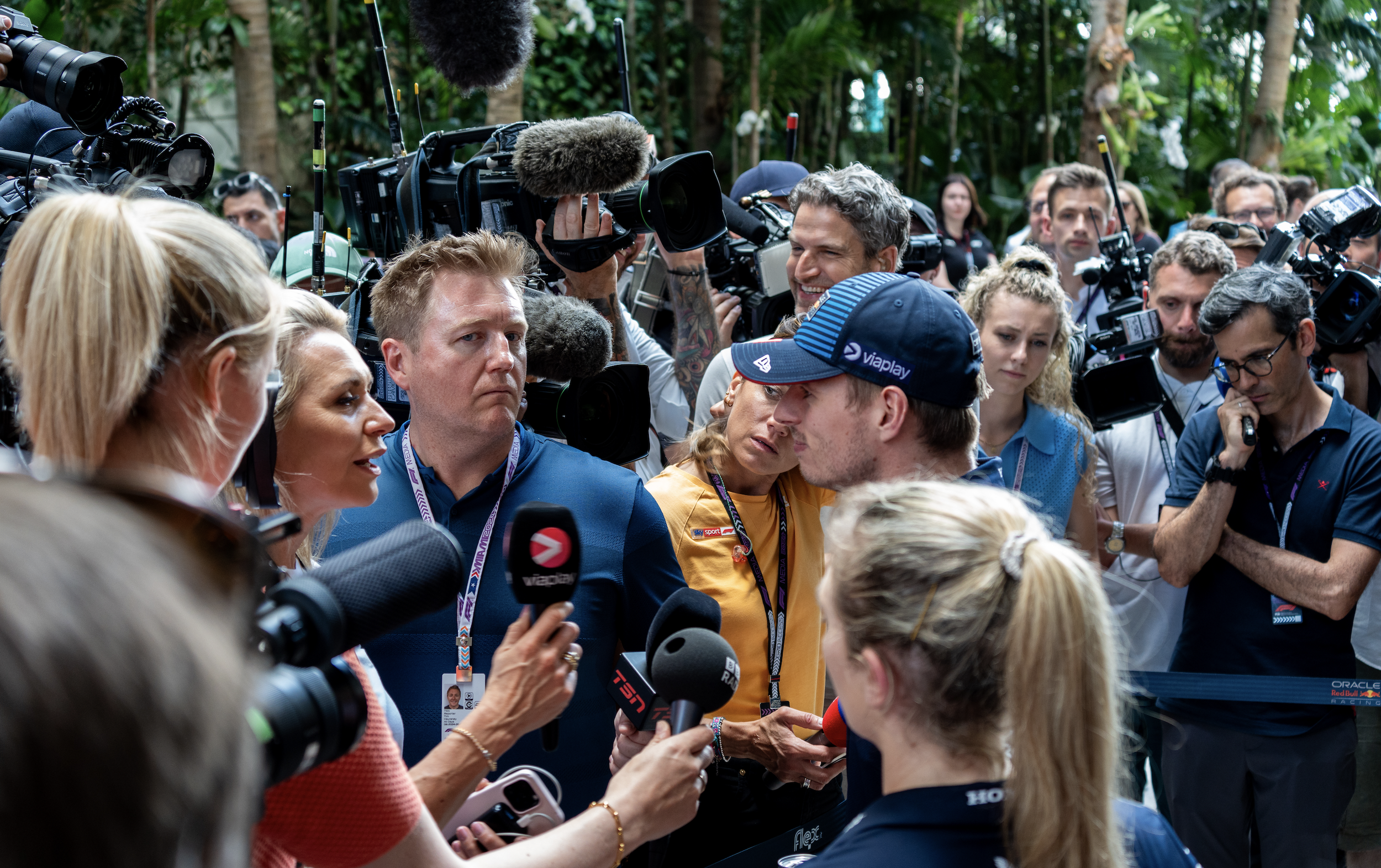 A group of journalists and photographers interview a man in a race suit, surrounded by cameras and microphones, outdoors with green trees in the background.