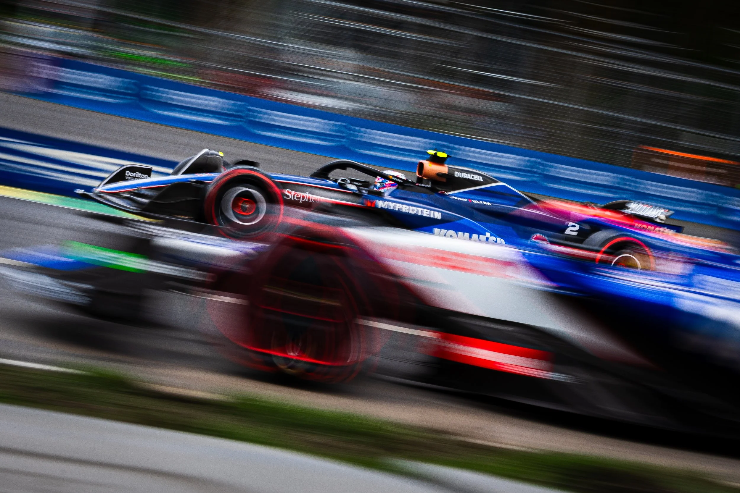 A race car speeding on a track with motion blur, featuring blue, black, and red colors, and various sponsor logos.