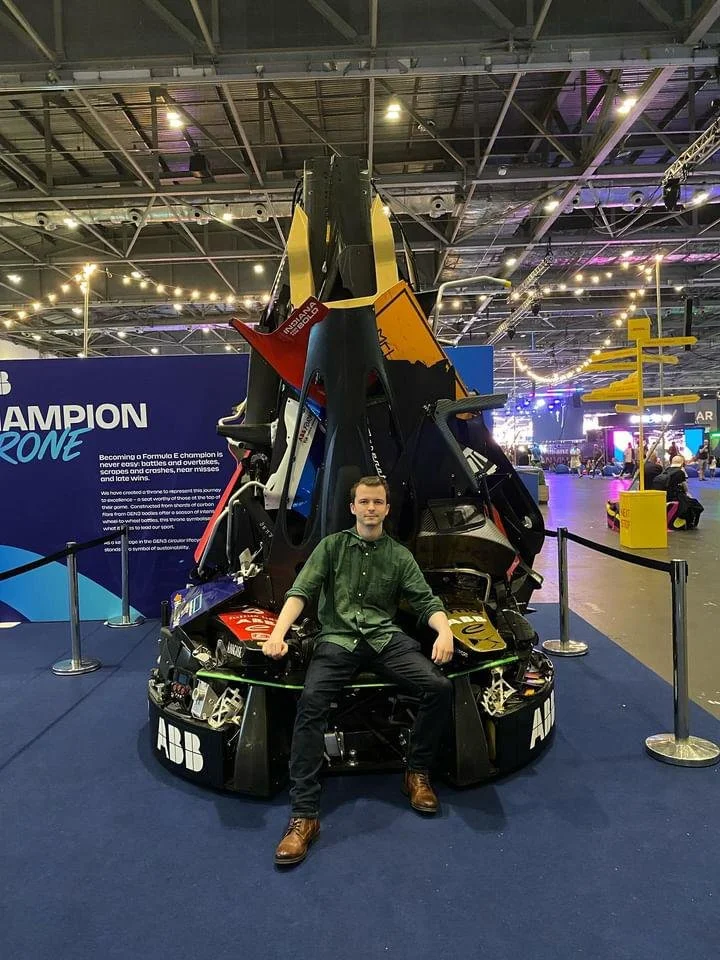 A young man sitting on a simulated cockpit or racing seat in front of a racing car, possibly an F1 or similar race car, at an indoor exhibition or tech event.