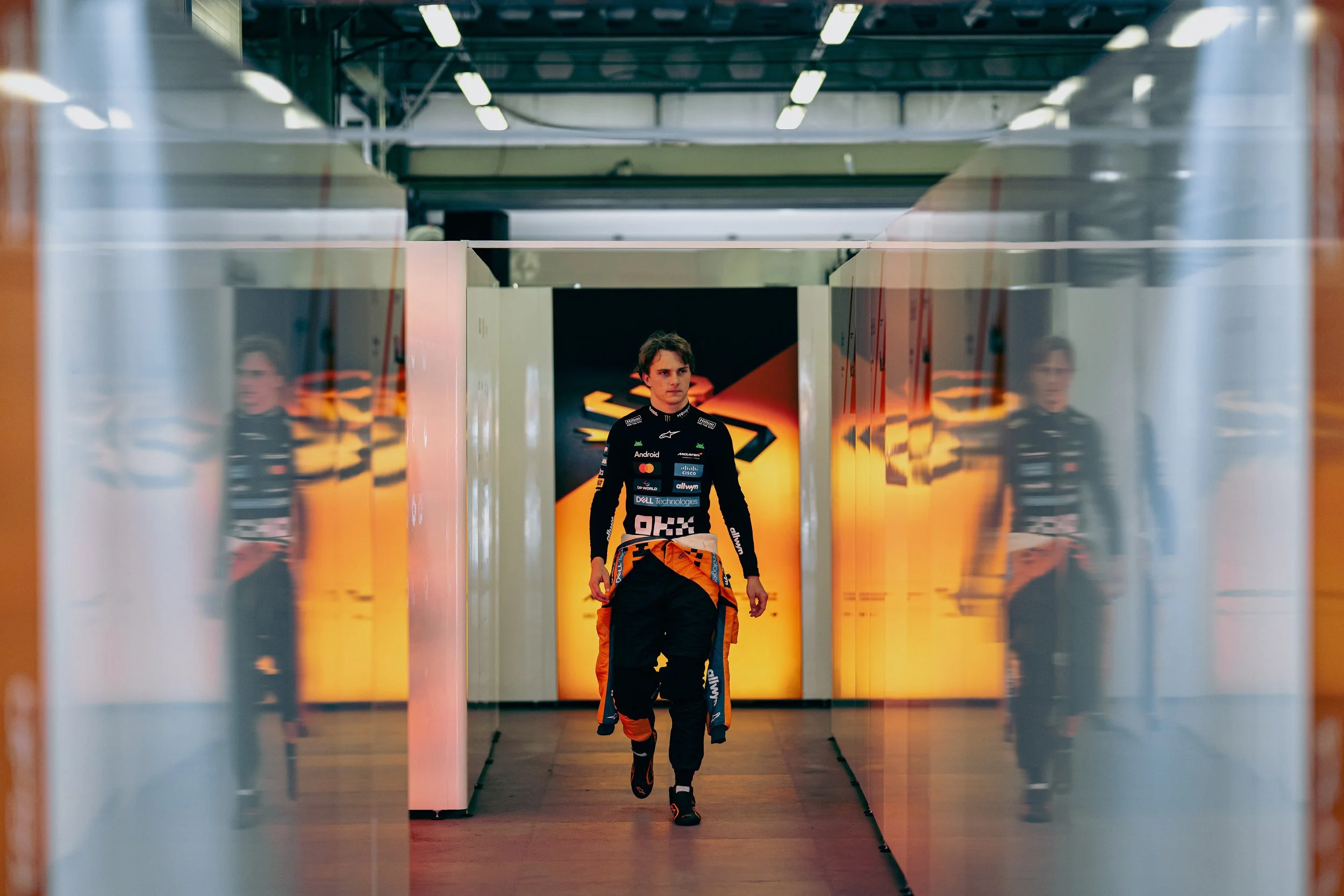 Oscar Piastri walks through a McLaren paddock corridor, reflected in the glass on either side.