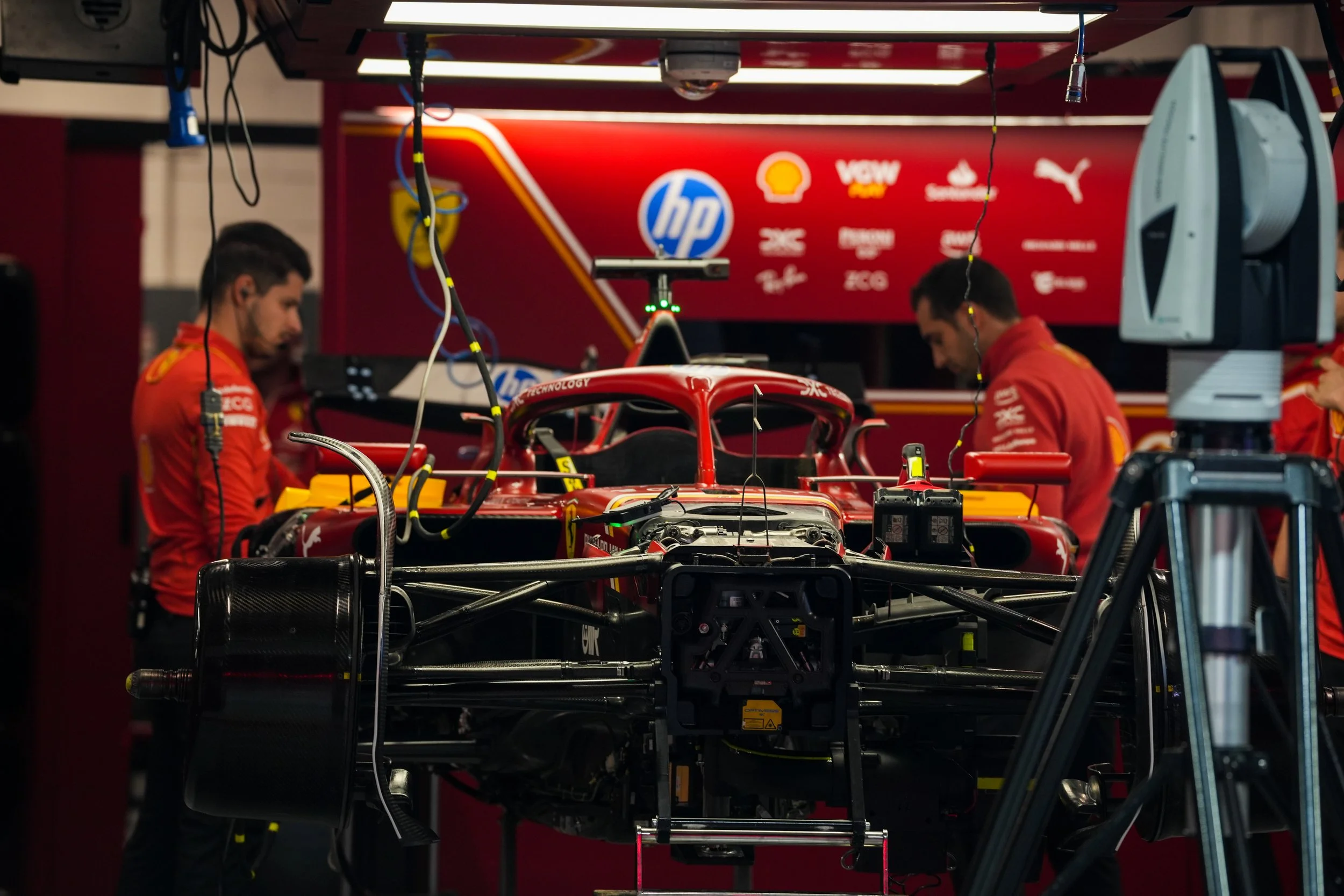 Scuderia Ferrari car in a garage with engineers working on it, surrounded by tools and equipment, with logos of sponsors in the background.