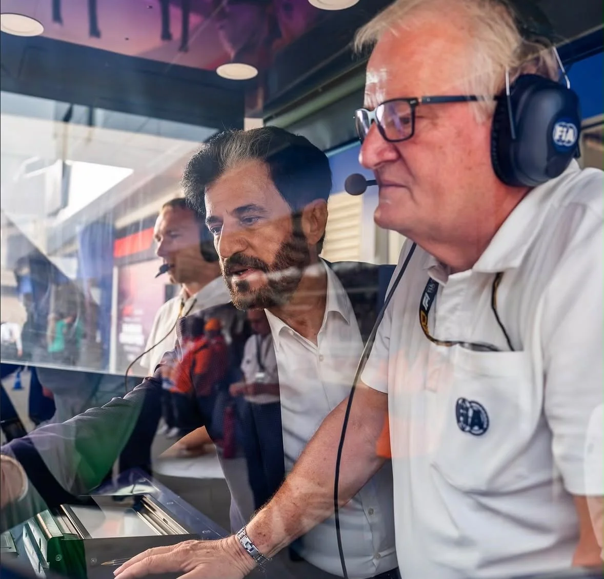Mohammed Ben Sulayem and an FIA official wearing a headset inside race control, photographed through reflective glass.