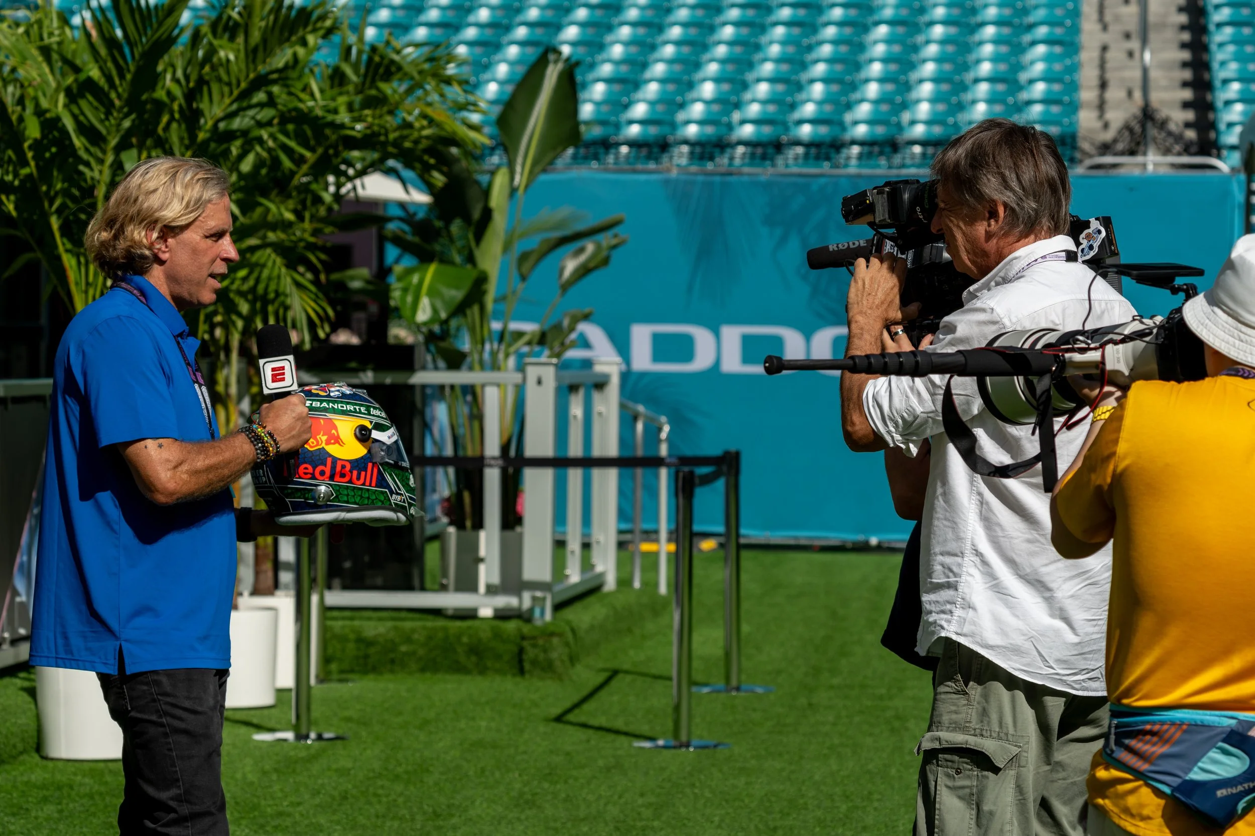 A man in a blue shirt holding a Red Bull racing helmet, being interviewed by a person with a camera at a race track.